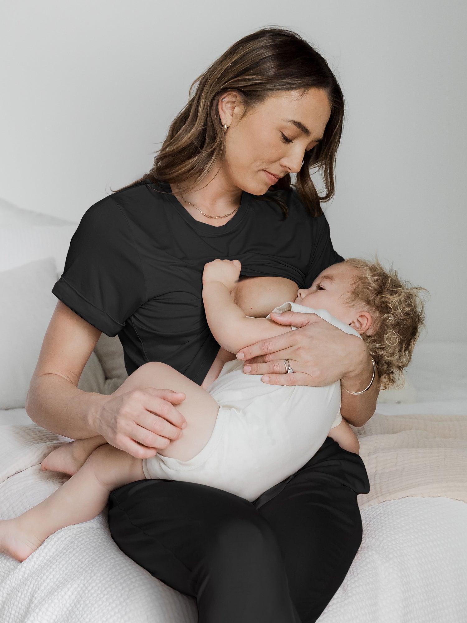 Model sitting on a bed wearing the Everyday Asymmetrical Nursing T-Shirt in Black breastfeeding her baby through the easy nursing access. @model_info:Kate is wearing a Small.