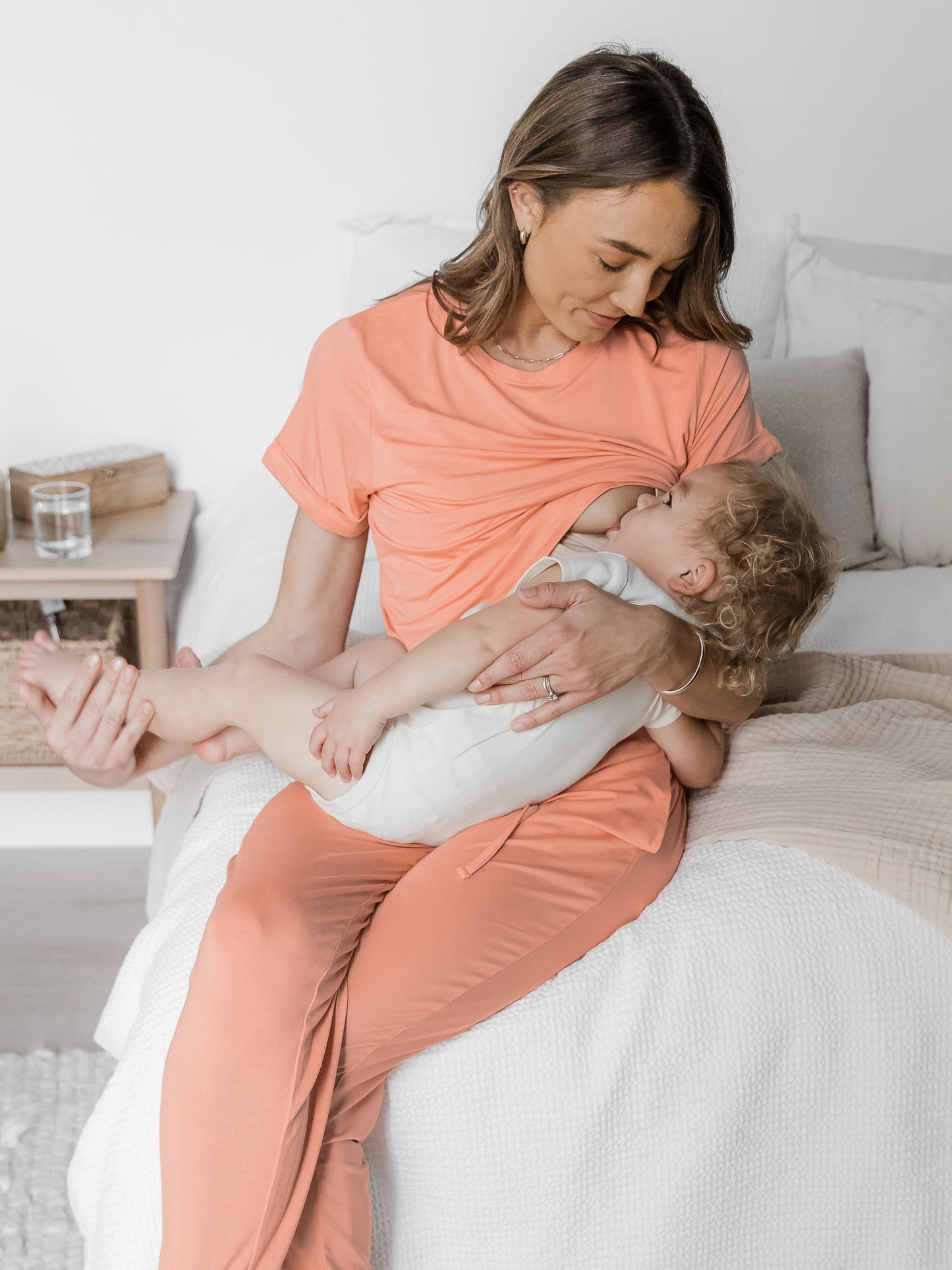 Breastfeeding model sitting on a bed with her baby while wearing the Everyday Symmetrical Nursing T-Shirt in Vintage Coral. @model_info:Kate is wearing a Small.