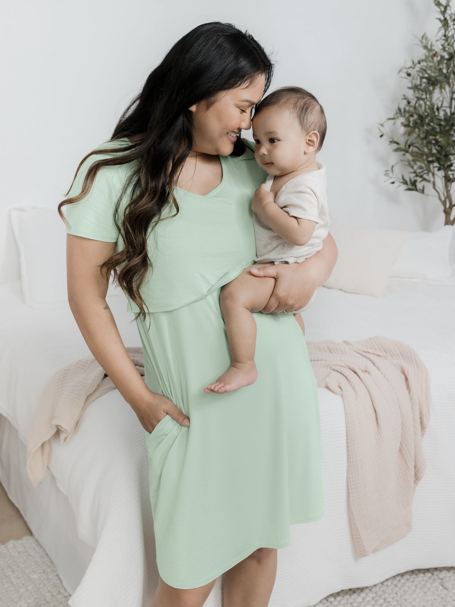 Model holding her baby on her hip standing in front of a bed and wearing the Eleanora Bamboo Maternity & Nursing Dress in Soft Mint. @model_info:Binc is 5'5" and wearing a Medium.