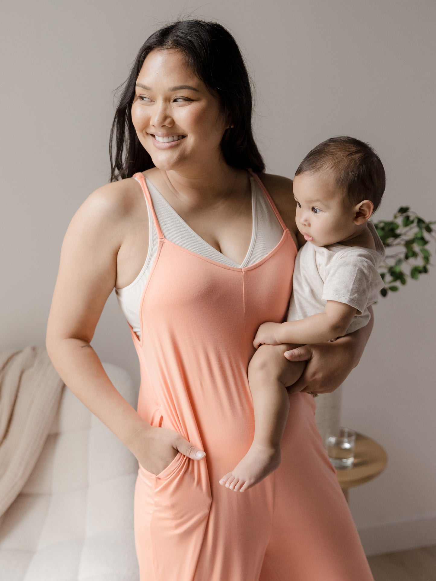 Model holding her baby with her hand in her pocket while wearing the Charlie Maternity & Nursing Romper in Vintage Coral. @model_info:Binc is 5'5" and wearing a Medium.