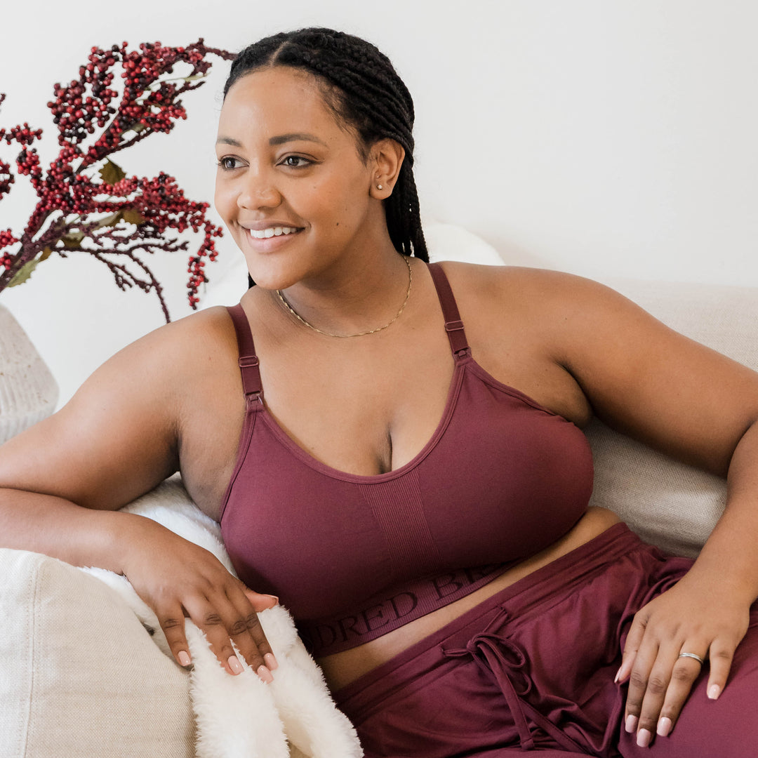 Model wearing the Sublime® Hands-Free Pumping & Nursing Sports Bra in Fig sitting on a couch. @model_info:Roxanne is wearing a Large.