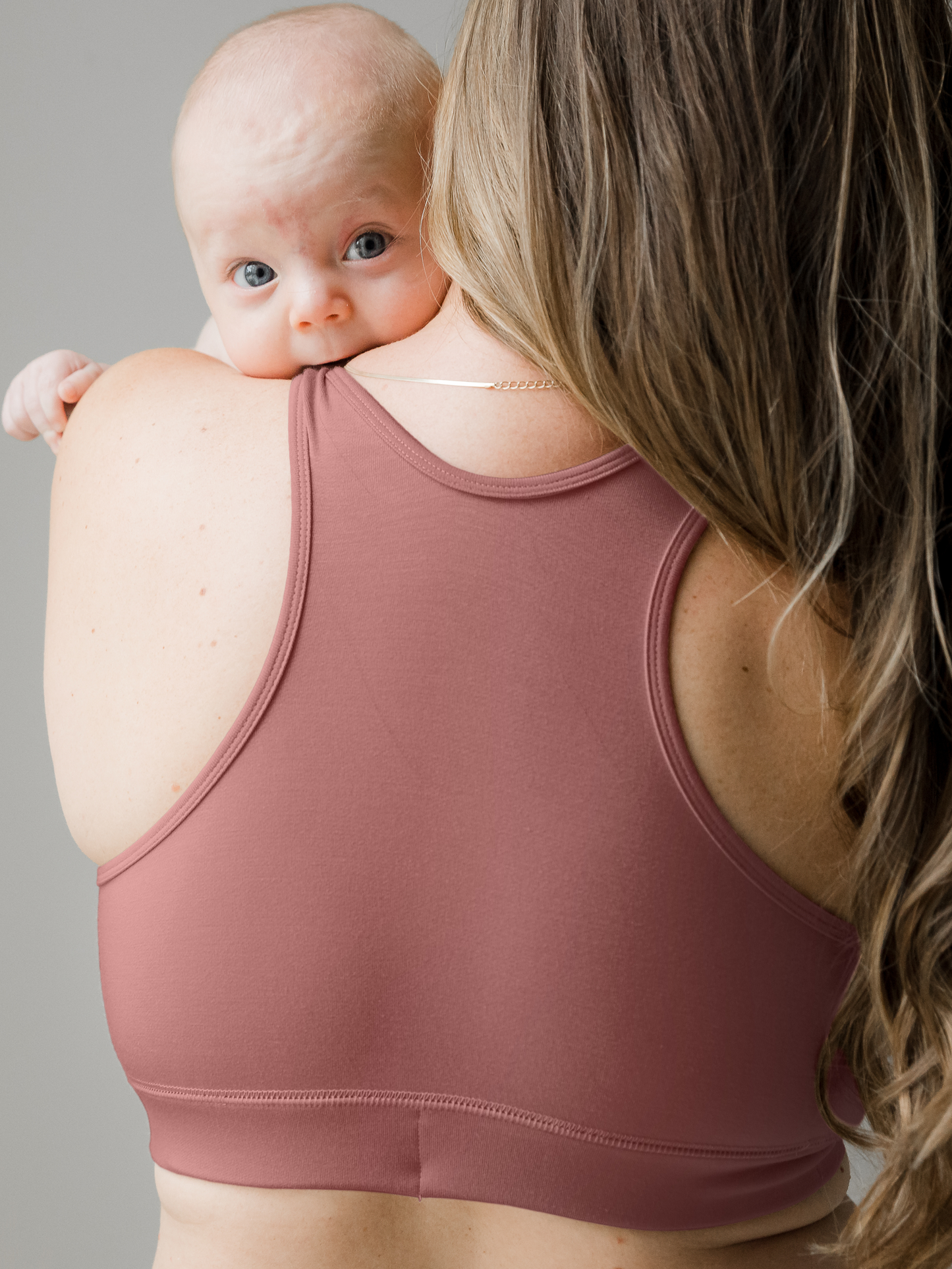 Back view of model wearing the French Terry Racerback Nursing & Sleep Bra in Redwood holding her baby who is peeking over her shoulder