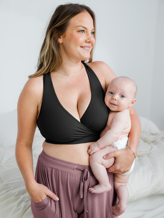 Model wearing the French Terry Racerback Nursing & Sleep Bra in Black holding her baby against her chest. @model_info:Ali is wearing a Large Busty.