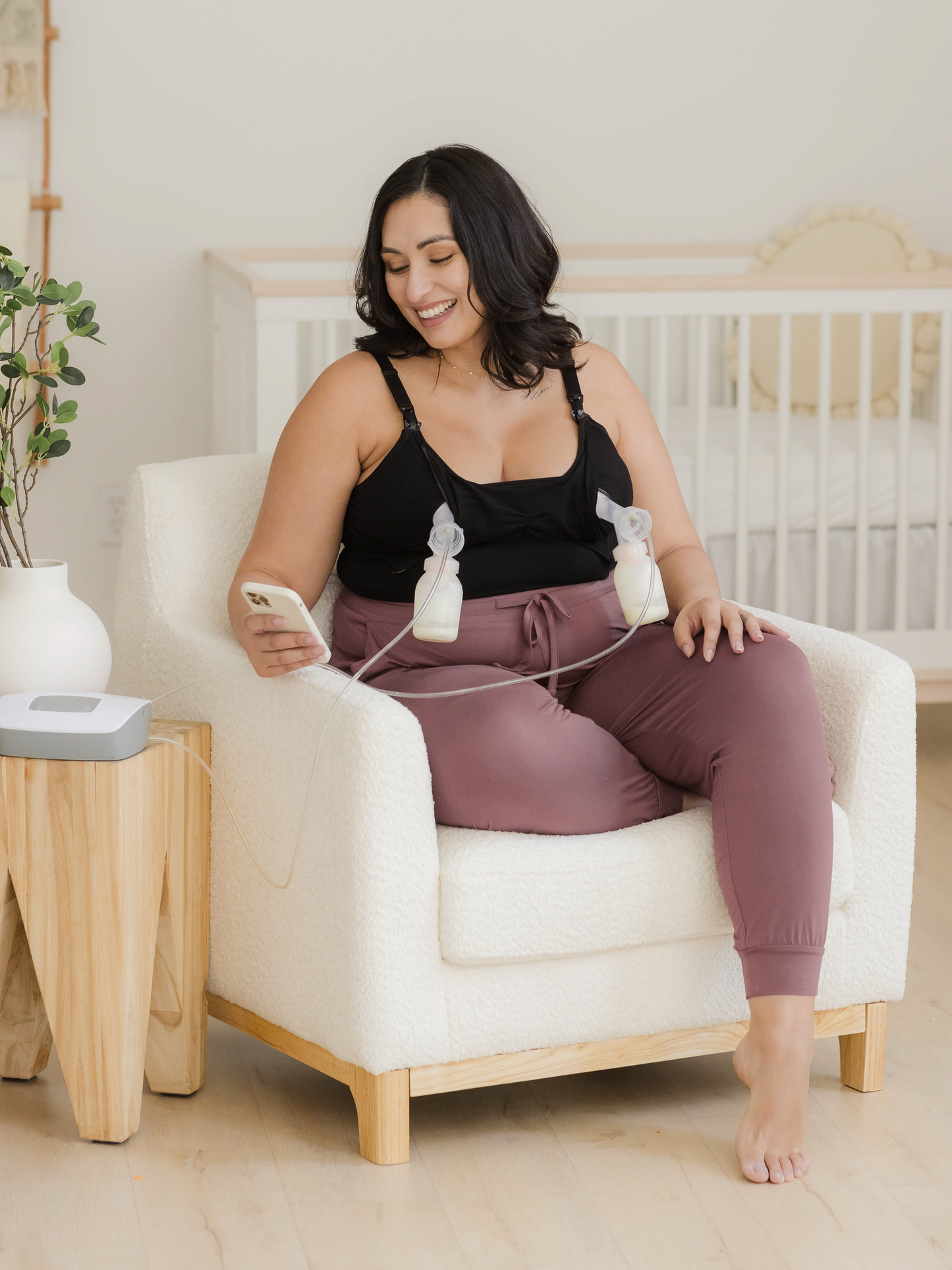 Model sitting in chair pumping and holding phone and wearing the Sublime® Bamboo Hands-Free Pumping & Nursing Camisole in Black@model_info:Connie is wearing an X-Large Busty.
