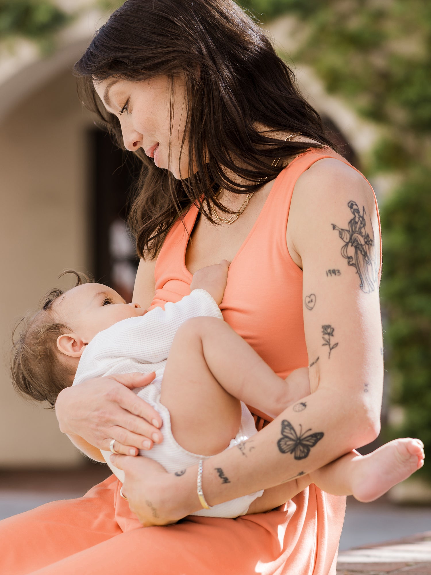 Model sitting and nursing baby wearing the Penelope Crossover Nursing Dress in vintage coral