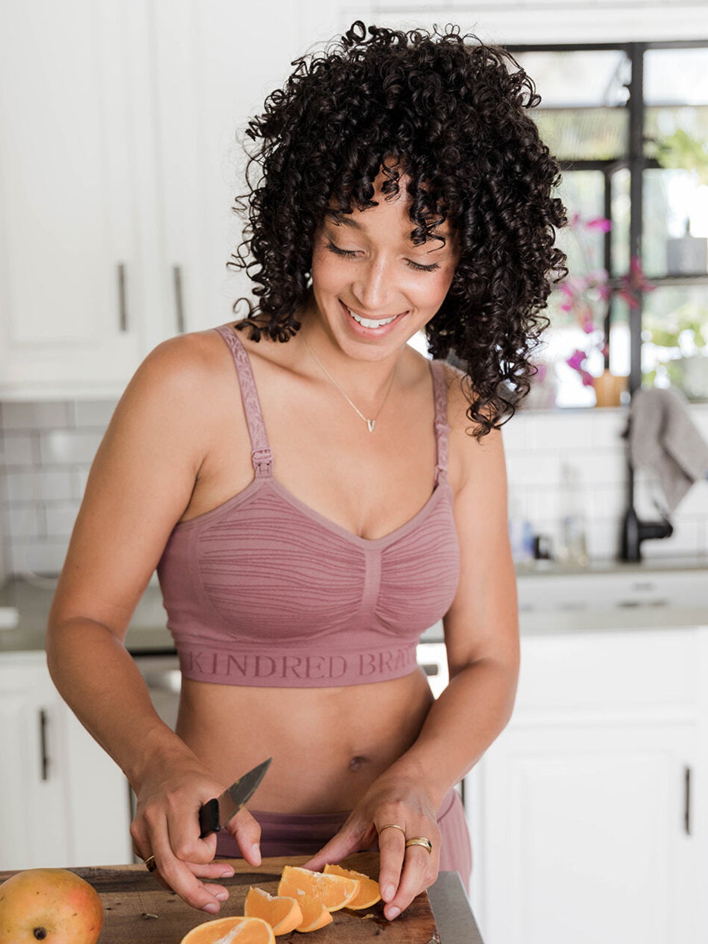 A model wearing the Sublime® Hands-Free Pumping & Nursing Bra in twilight chopping oranges in the kitchen @model_info:Arion is wearing a Small. 