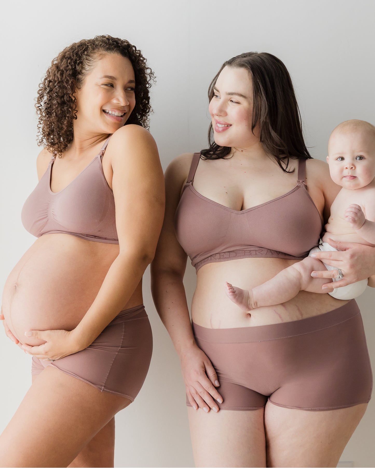 One model is pregnant, holding bump and other model is holding her baby, both smiling and wearing Grow with Me™ Maternity & Postpartum Boyshort in soft pink on a beige background.