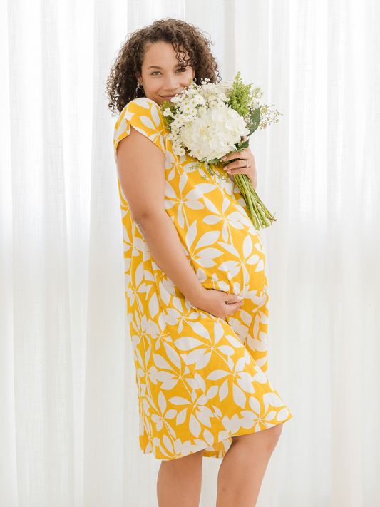 Side view of pregnant model wearing the Universal Labor & Delivery Gown in Honey Leaf, with one hand under stomach and the other holding flowers up to face.@model_info:Alysha is 5'6" and wearing a S/M/L.