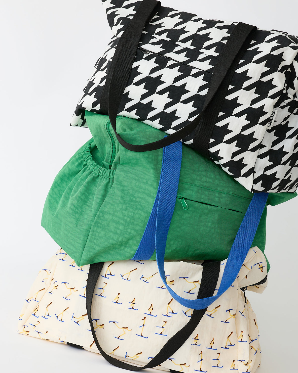 Three Large Weekender Bags are stacked on one another, Black Houndstooth, Green and Ivory Ski, on an off white background.