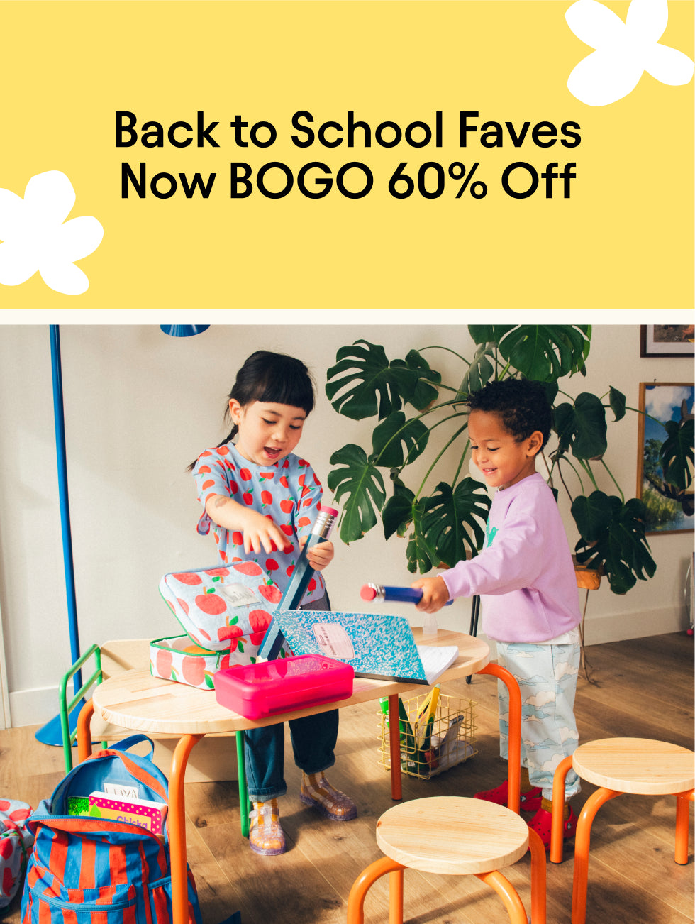 Promotional image with yellow background and white flower shapes and black text reading, Back to School Faves Now BOGO 60% Off. Image with two children playing in a play space with table, chairs, notebooks, oversized pencils, Cool Kid Backpacks, Cool Kid Lunch Boxes and Graphic Tee in Blue Apple.