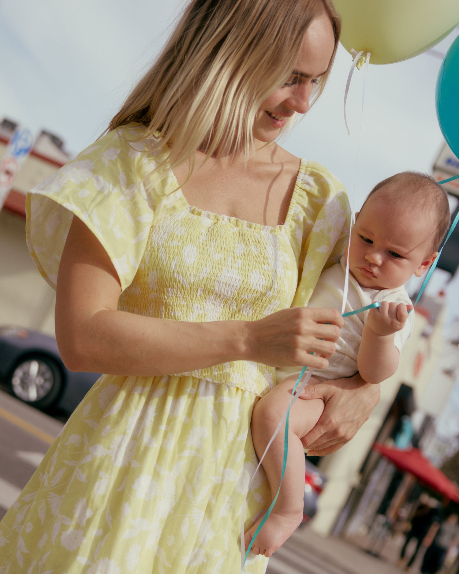 Woman in the Lola Smocked Maternity & Nursing Midi Dress in a Yellow Floral print holding a baby with balloons in an outdoor setting