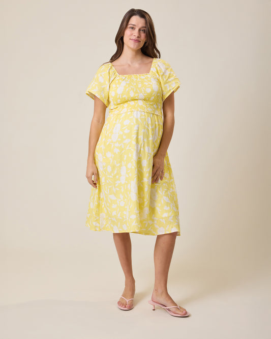 Woman wearing the Lola Smocked Maternity & Nursing Midi Dress in a Yellow Floral print with a beige background @model_info:Elliana is 5'7" and wearing a Medium.