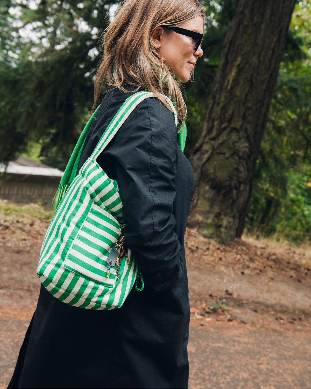 Model is walking outdoors in a natural park setting, while wearing sunglasses, long black coat, green scarf and Stroller Party™ Bucket Bag in Green Stripe. 