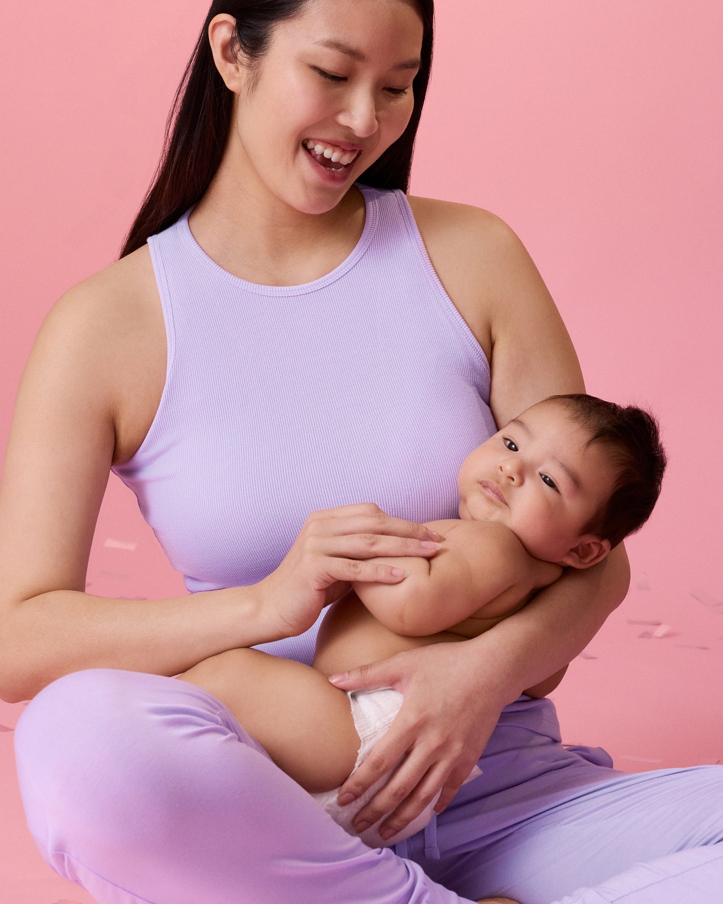 Model wearing Ribbed Bamboo Racerback Nursing Tank in Lavender sitting with crossed legs and smiling at the baby in her arms
