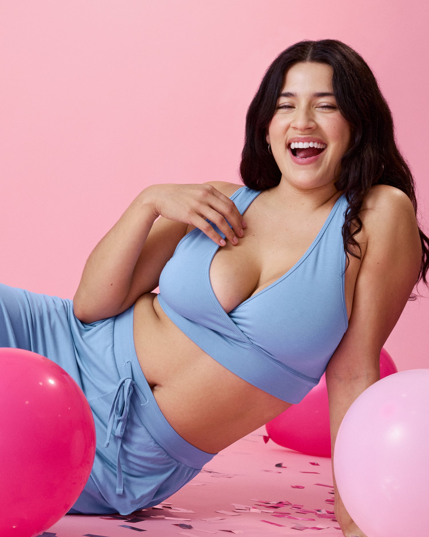 Model wearing French Terry Racerback Nursing & Sleep Bra in Vintage Blue on a pink background sitting on the floor in a relaxed pose and surrounded by pink balloons and confetti 