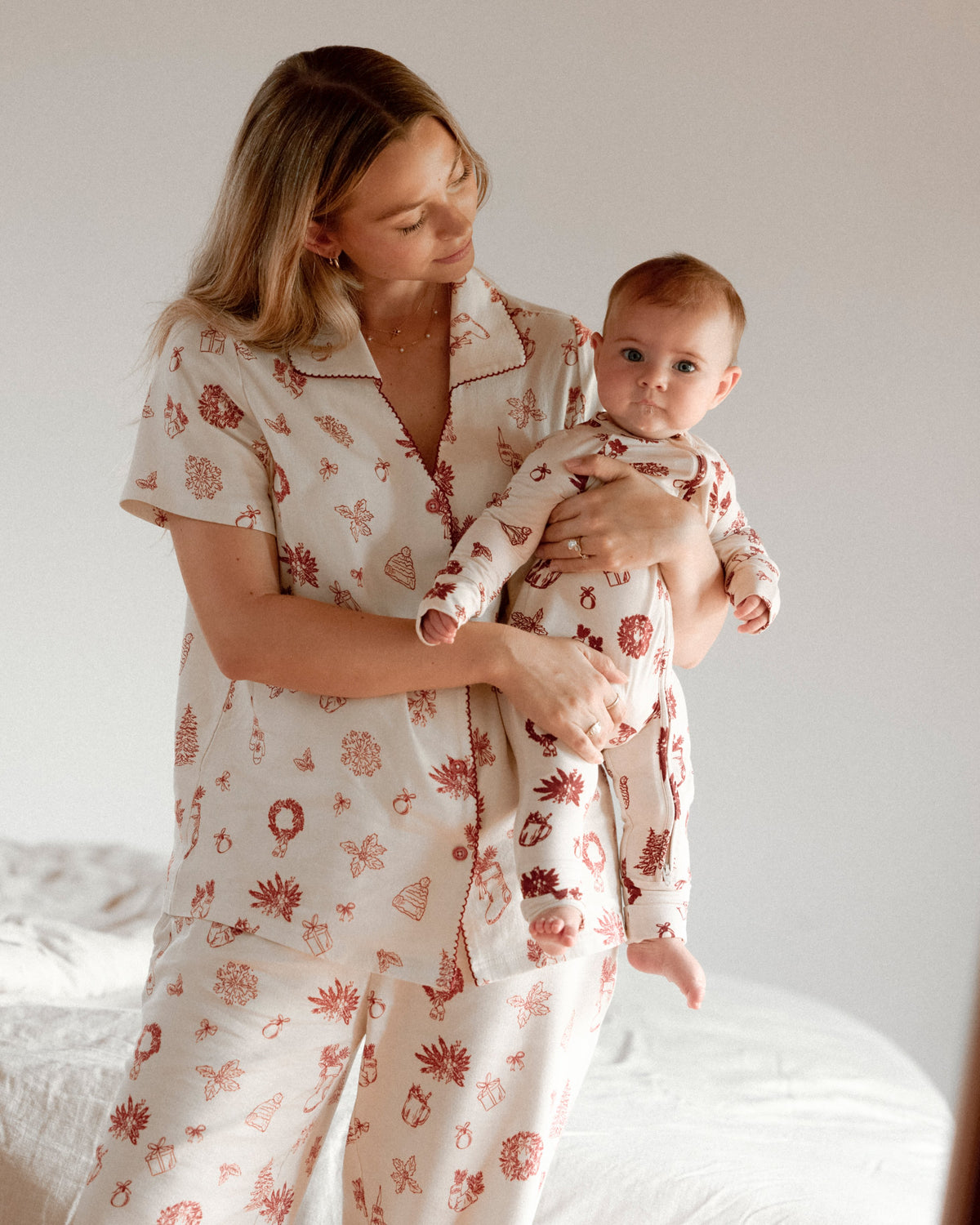 A woman and baby wearing matching holiday-themed pajamas standing by a bed. Mother is wearing Cotton Bamboo Pajama Set in Cozy Day and baby is wearing Bamboo Baby Pajama in Cozy Day. In a sun lit bedroom. @model_info:Saige is 5'7" and wearing a Small.
