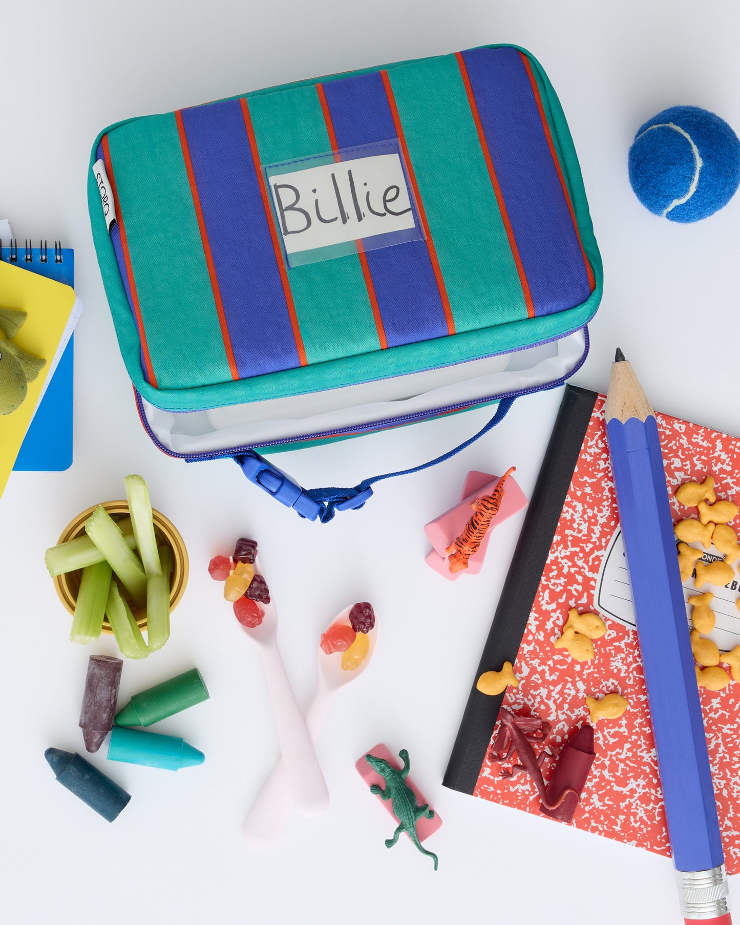 Flat lay of Cool Kid Lunch Box in Navy & Green Stripe with giant pencil, tennis ball, notebooks, snacks, crayons, goldfish, miniature animals and erasers on a beige background.