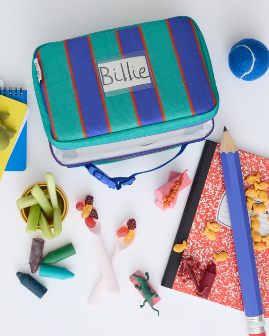Flat lay of Cool Kid Lunch Box in Navy & Green Stripe with giant pencil, tennis ball, notebooks, snacks, crayons, goldfish, miniature animals and erasers on a beige background.