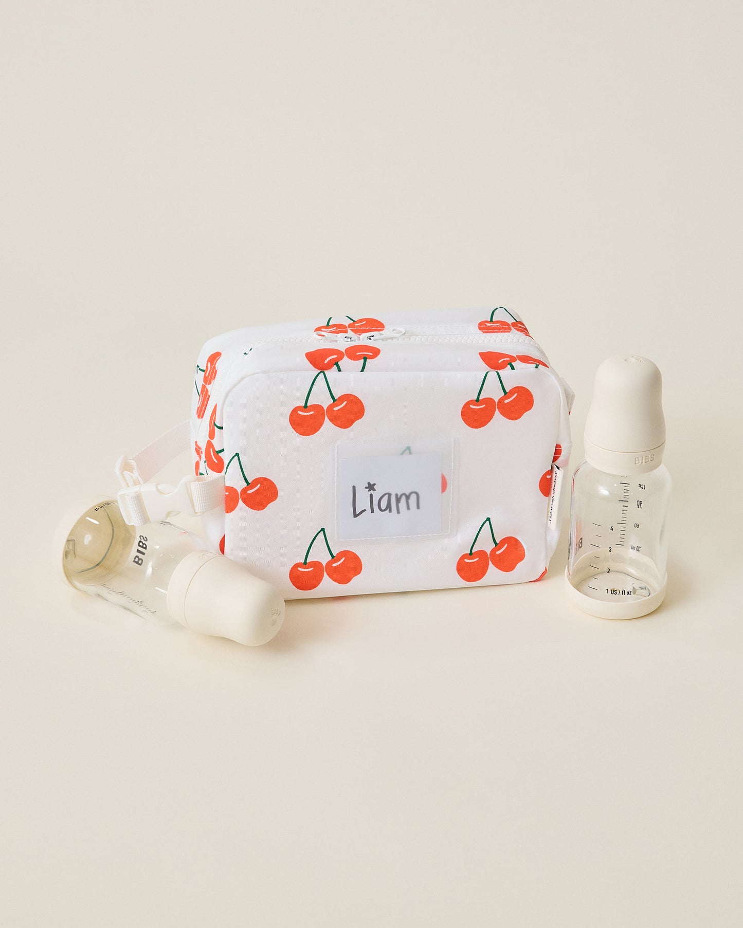 White Mini Cooler Bag with red cherry pattern, sitting next to two bottles, on a beige background