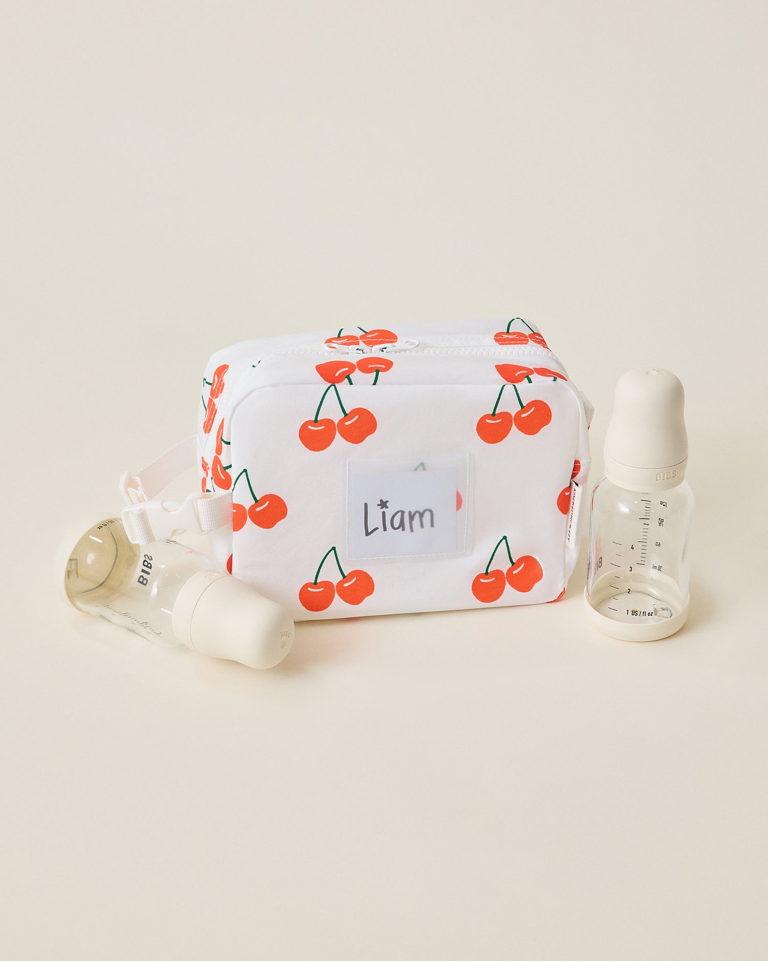 White Mini Cooler Bag with red cherry pattern, sitting next to two bottles, on a beige background