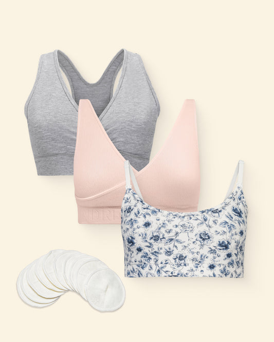 The Mix-and-Match Bralette Bundle on a beige background. French Terry Maternity & Nursing Bralette in navy toile blue floral pattern, French Terry Racerback Nursing & Sleep Bra in grey heather, Model is wearing Model wearing the Sublime® Adjustable Crossover Nursing & Lounge Bra in Soft Pink and ten Organic Bamboo Reusable Nursing Pads. 
