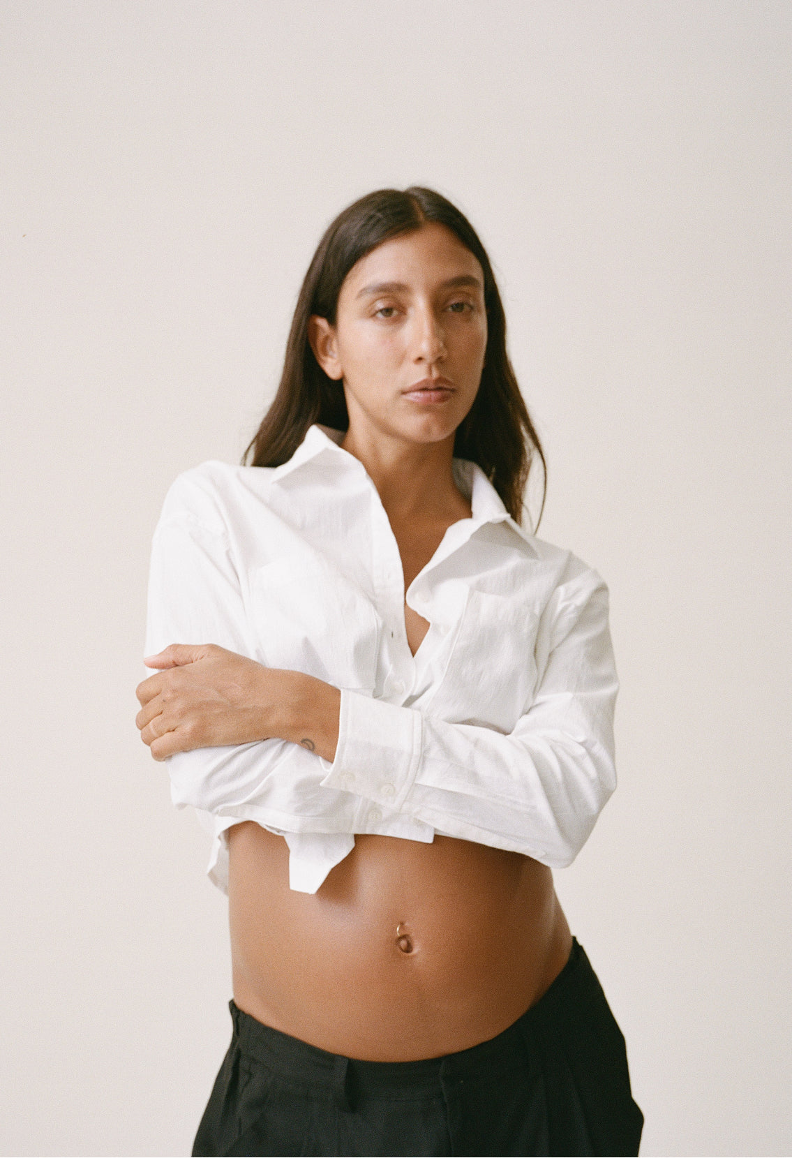 Pregnant woman wearing the white Poplin Maternity & Nursing Button Up Shirt with button buttons undone, with black pants, against a solid beige background.