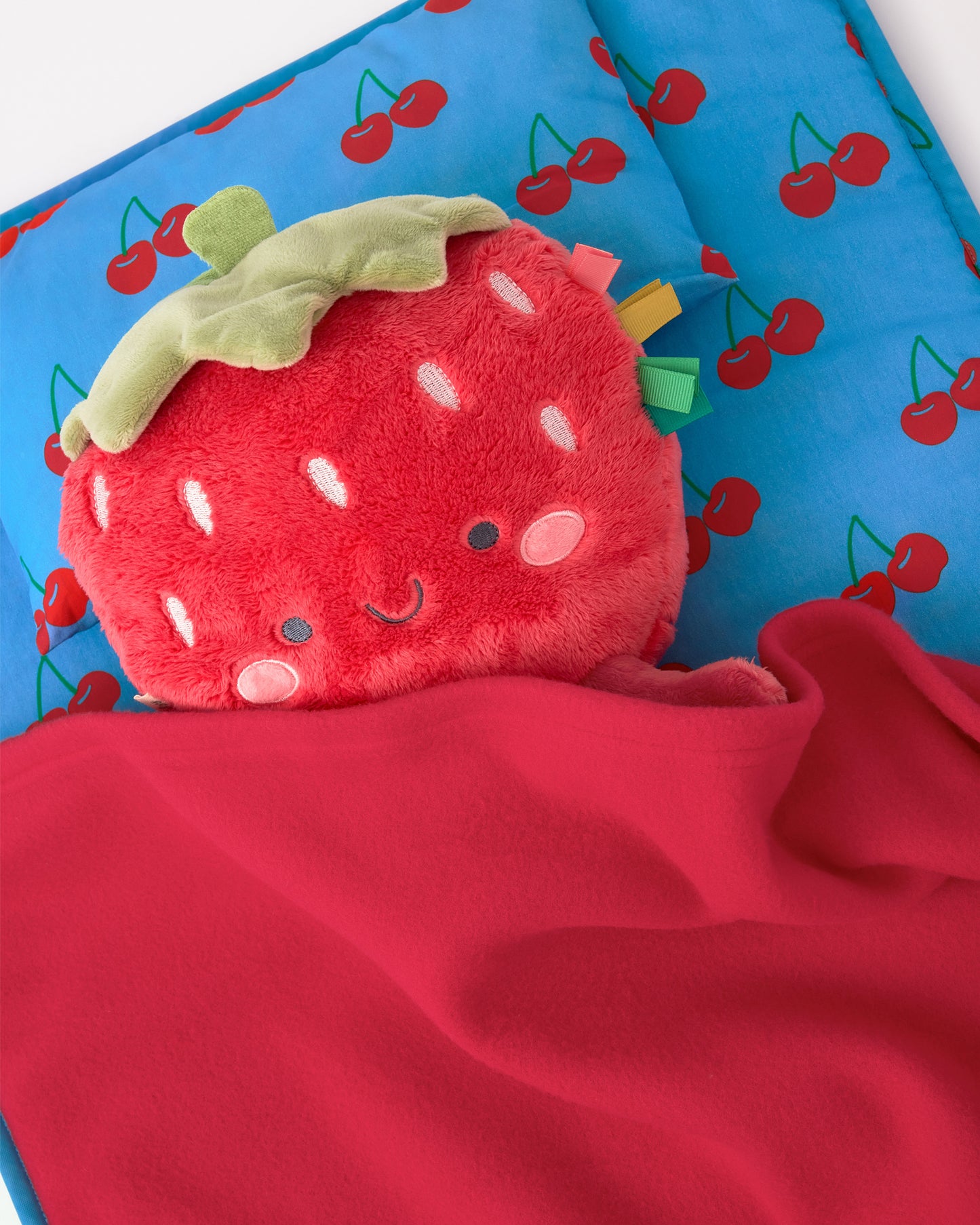Close up of the Nap Mat in Blue Cherries pattern with a strawberry doll on the pillow, on a beige background.