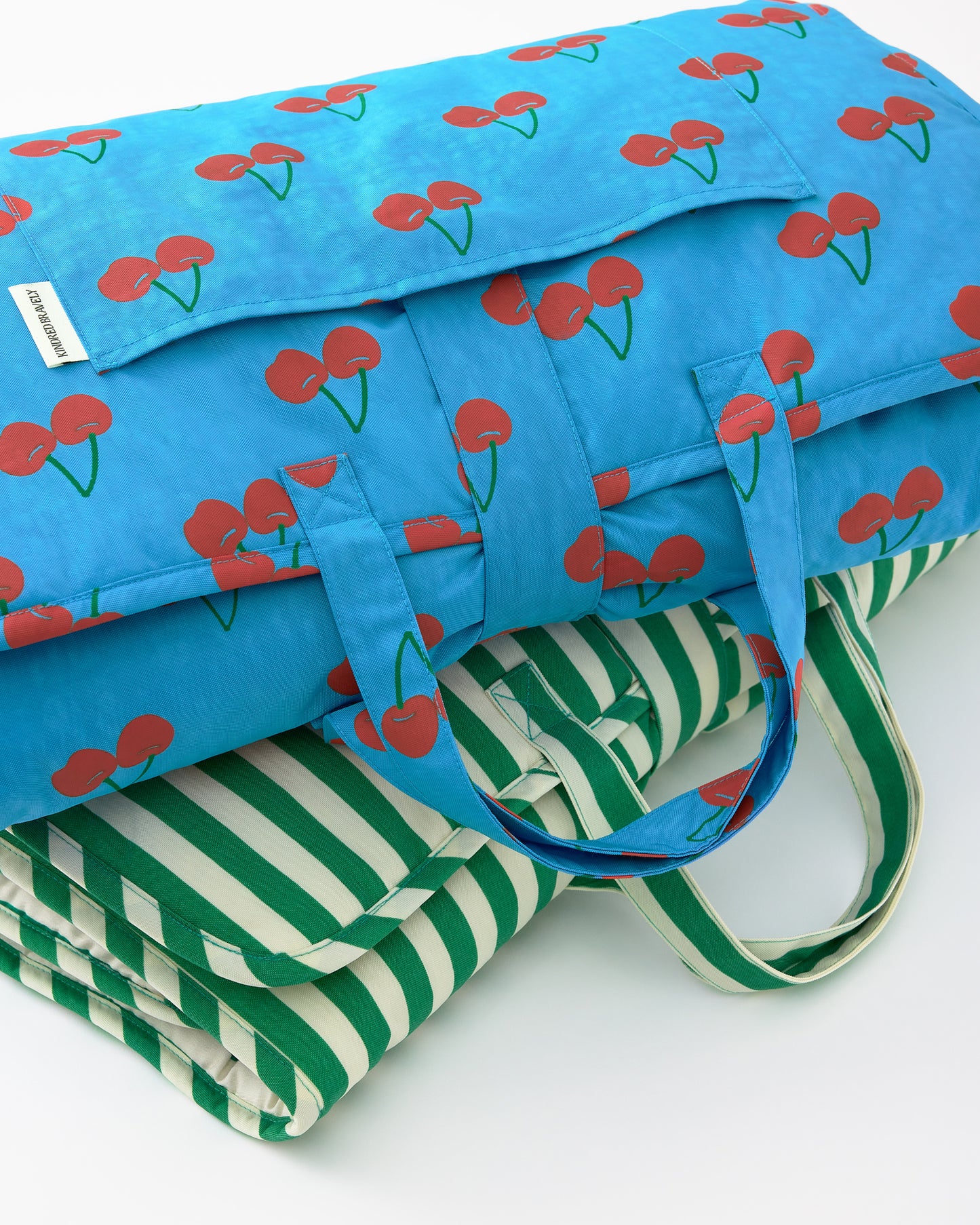 Close up of the folded up view of the Nap Mats in Blue Cherries and Green Stripe patterns on a beige background.