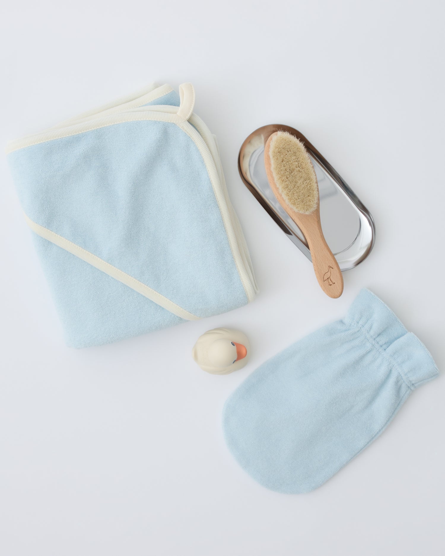 Newborn Bath Set in Breeze, including hooded towel, hair brush and bath mitt with brush on metallic soap dish next to rubber ducky, on a beige background.