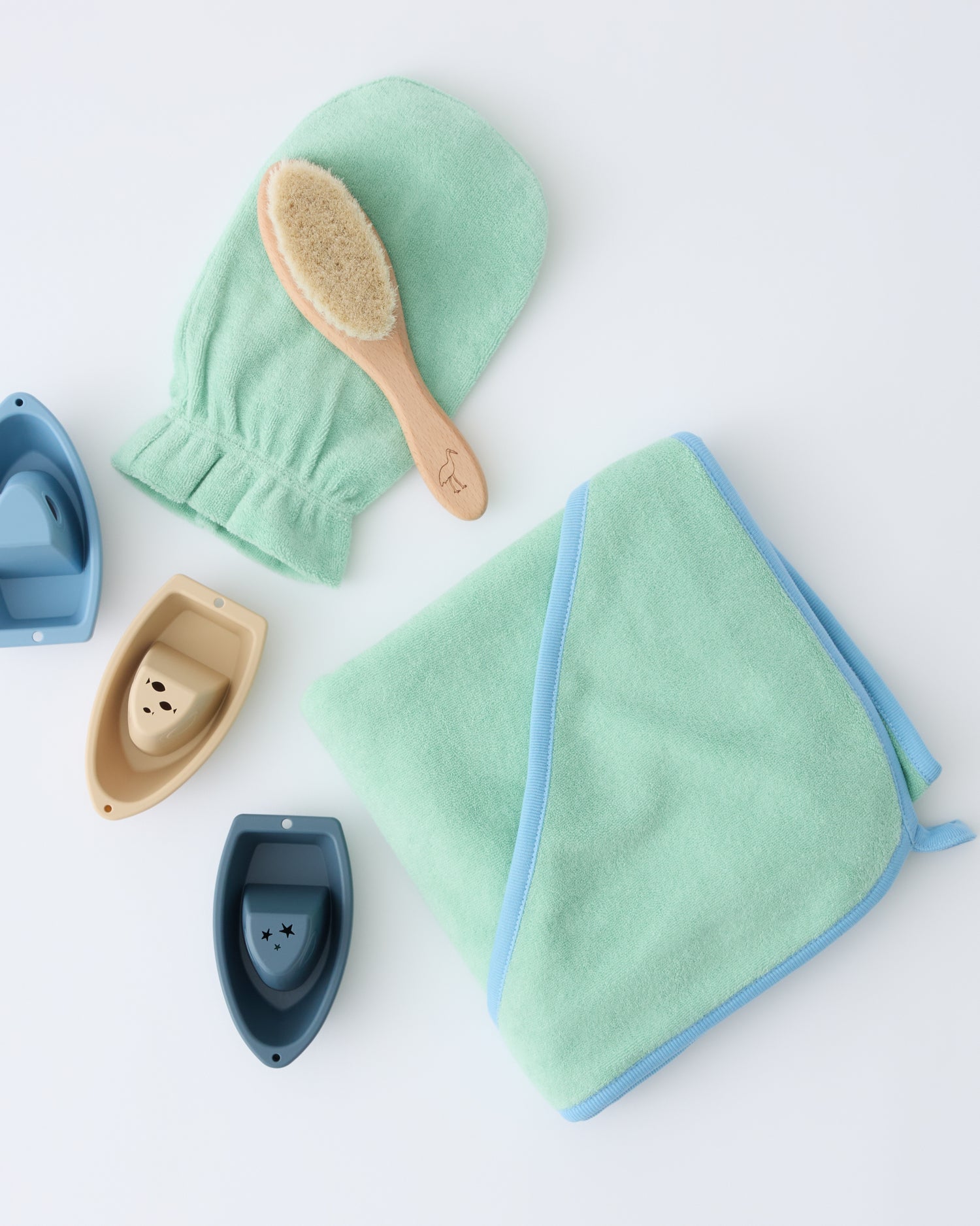 Newborn Bath Set in Mint, including hooded towel, hair brush and bath mitt, paired with three floating boat toys, on a beige background.