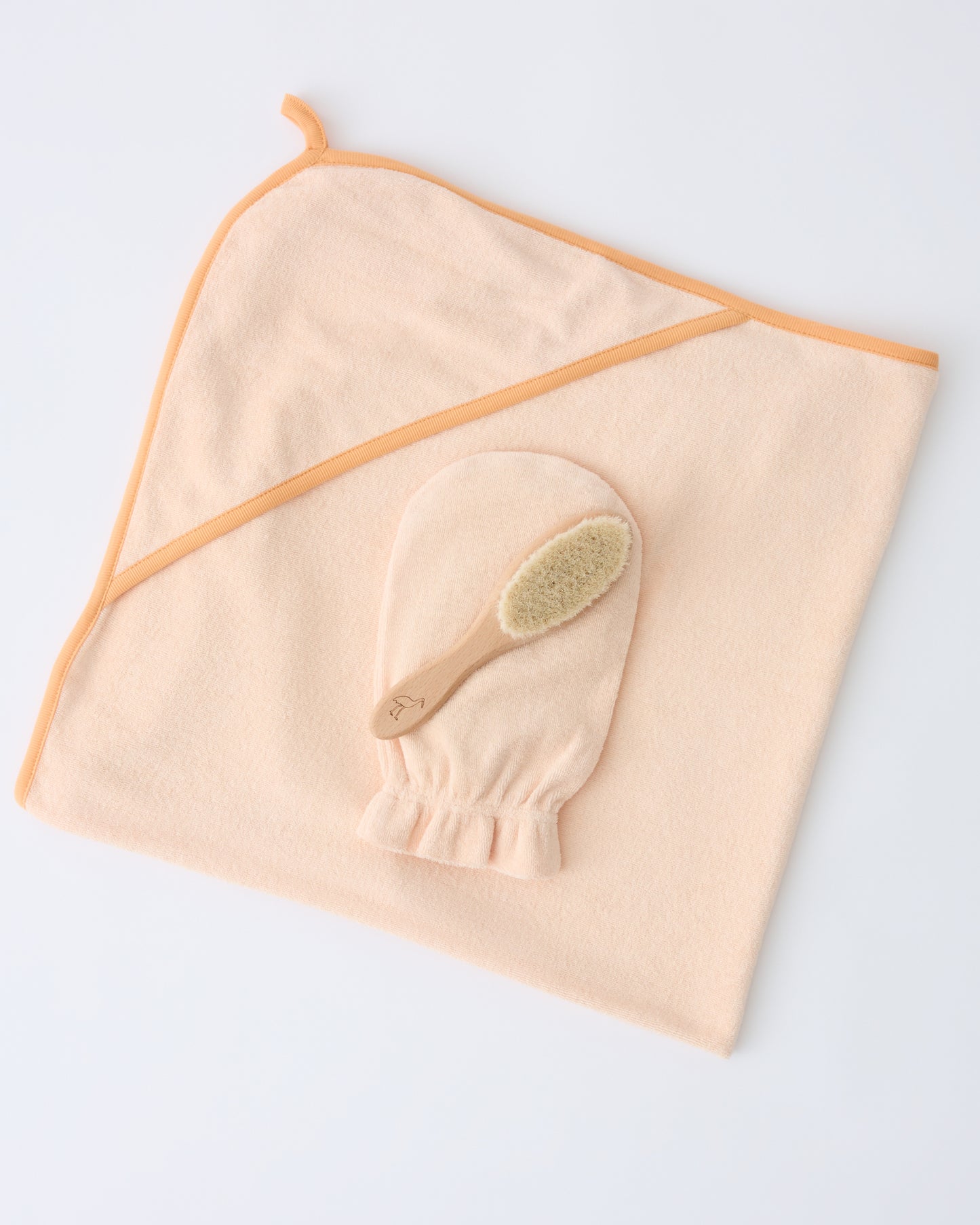 Newborn Bath Set in Peach Whip, including hooded towel, hair brush and bath mitt on a beige background.