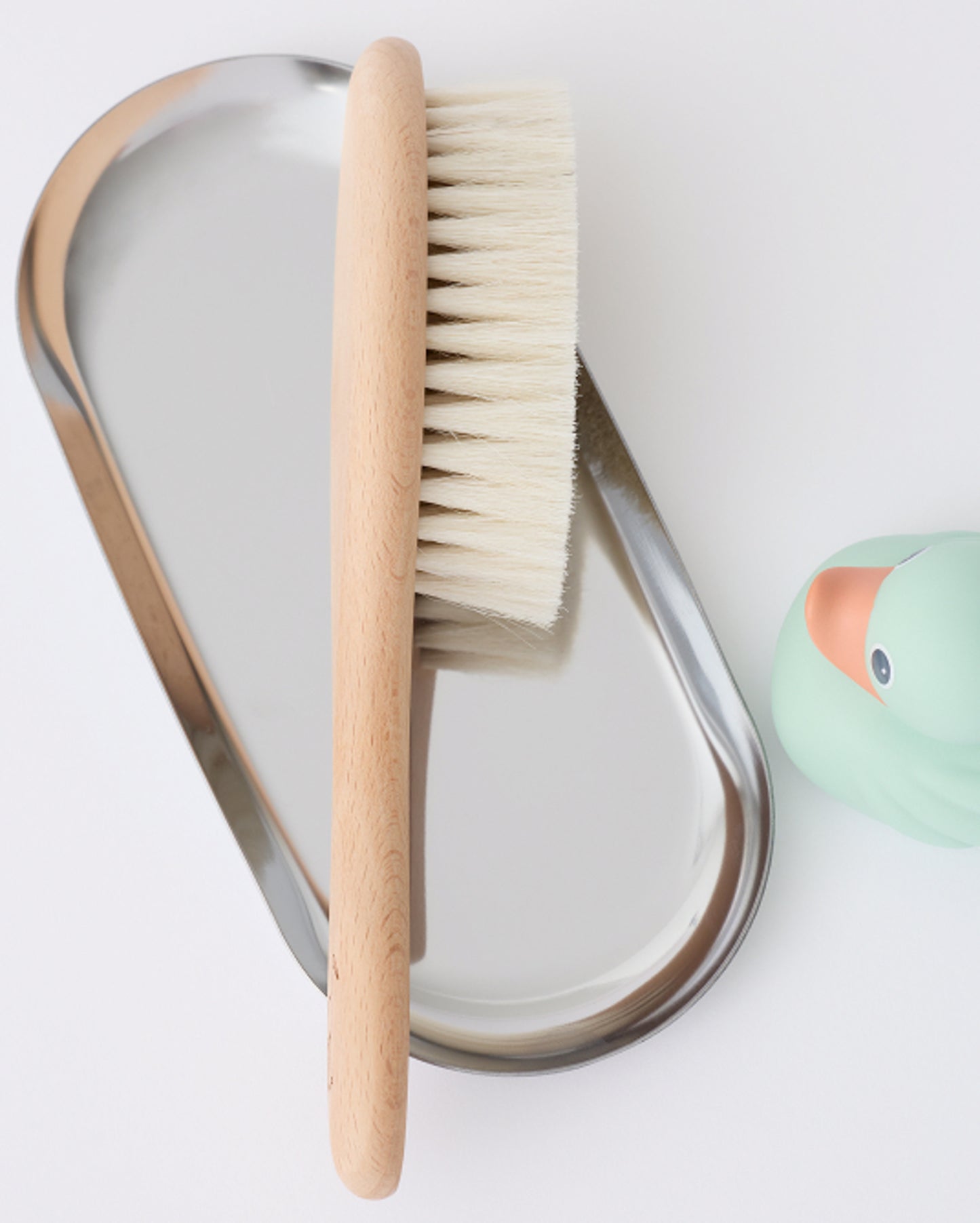 Natural wood baby hairbrush from the Newborn Bath Set in Peach Whip, resting on metallic soap dish next to a rubber ducky, on a beige background.