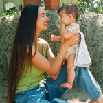 Woman holding baby outside, wearing a Pink Pomelo Onesie, with a Blue Wave Organic Muslin Burp Cloth, against a lush background.