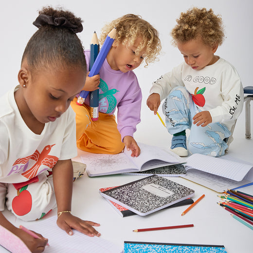 Three young children sitting on a white floor, surrounded by notebooks, colored pencils, and paper. The child on the left is writing and wearing Graphic Tee in White Bird, the child in the middle holds blue and purple colored pencils while wearing Graphic Sweatshirt in Purple Bird, and the child on the right looks at an open notebook while wearing Graphic Sweatshirt in Ivory Apple on a beige background.