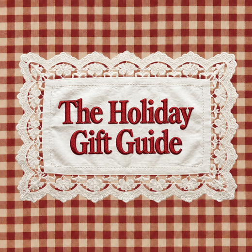 A square graphic with the text 'The Holiday Gift Guide' in red serif lettering on a white fabric-like panel with a scalloped lace border, set against a red and beige gingham check background.