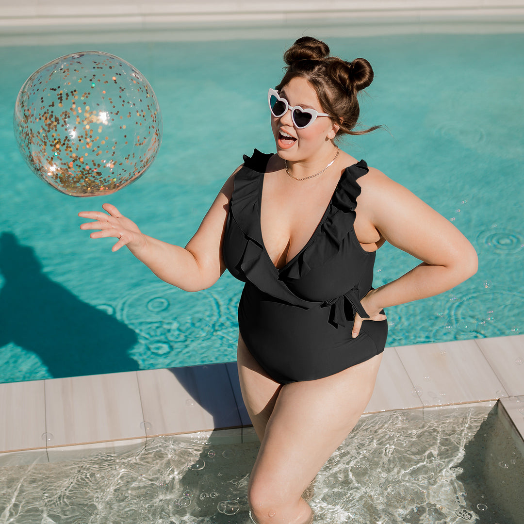 Model throwing a beachball up in the air while wearing the Ruffle Wrap Maternity & Nursing One Piece Swimsuit in Black