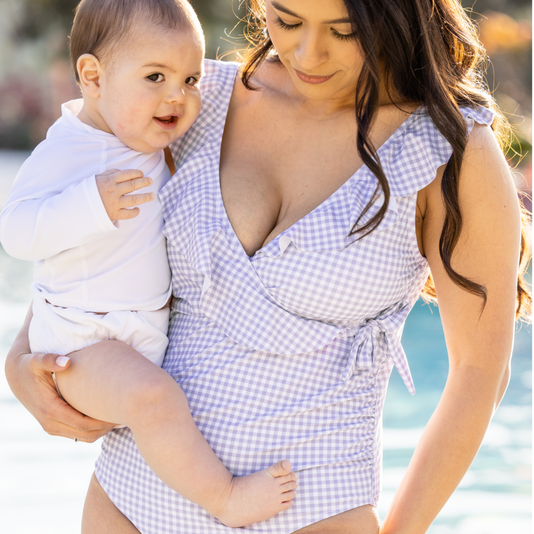 Model wearing the Ruffle Wrap Maternity & Nursing One Piece Swimsuit in Lavender Gingham with her baby on her hip standing by the pool. 