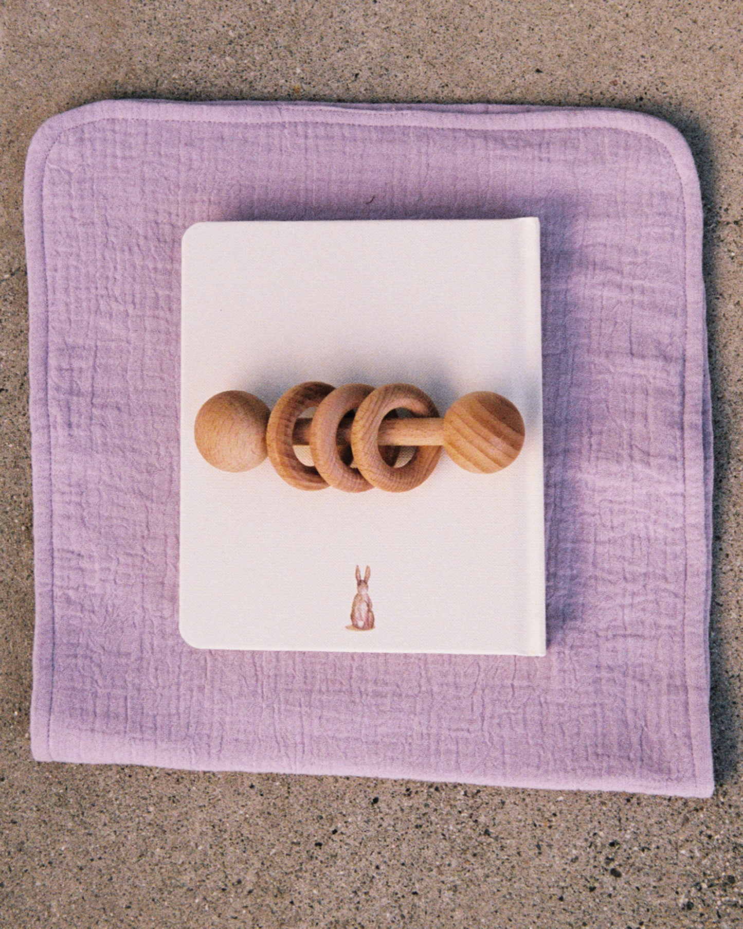 Organic Muslin Burp Cloth in the Lavender Cosmos print, displayed under a notebook and a wooden toy on a cement surface.