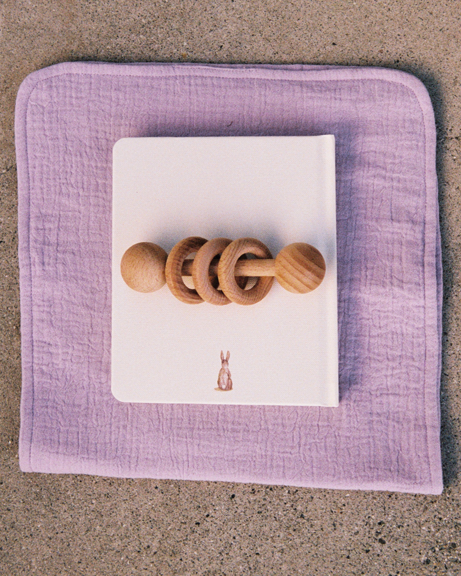 Organic Muslin Burp Cloth in the Lavender Cosmos print, displayed under a notebook and a wooden toy on a cement surface.