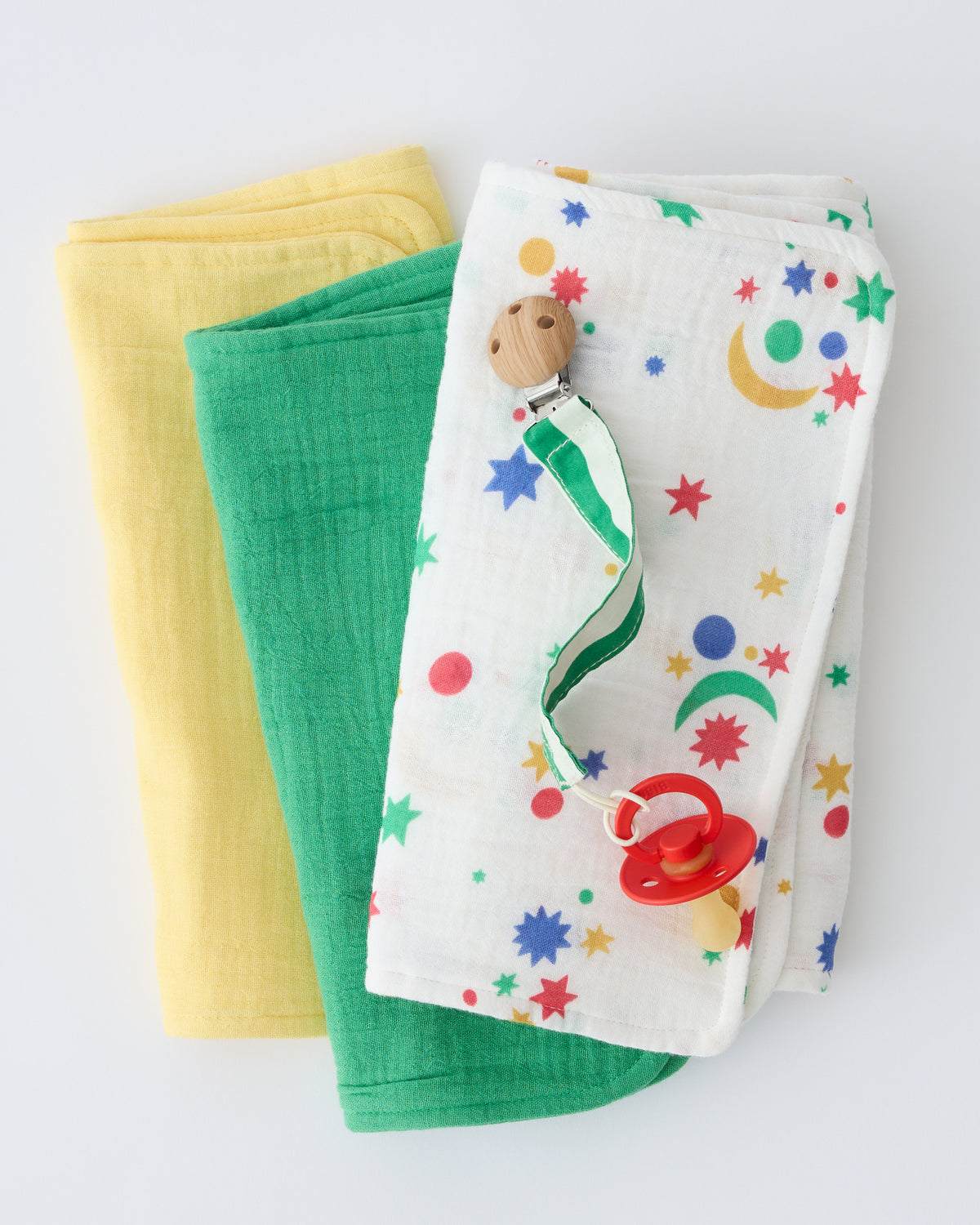 Organic Muslin Burp Cloth (Three-Pack) in White Starry Sky pattern, folded and stacked, with Pacifier Clip in Green Stripe ontop, on a beige background.