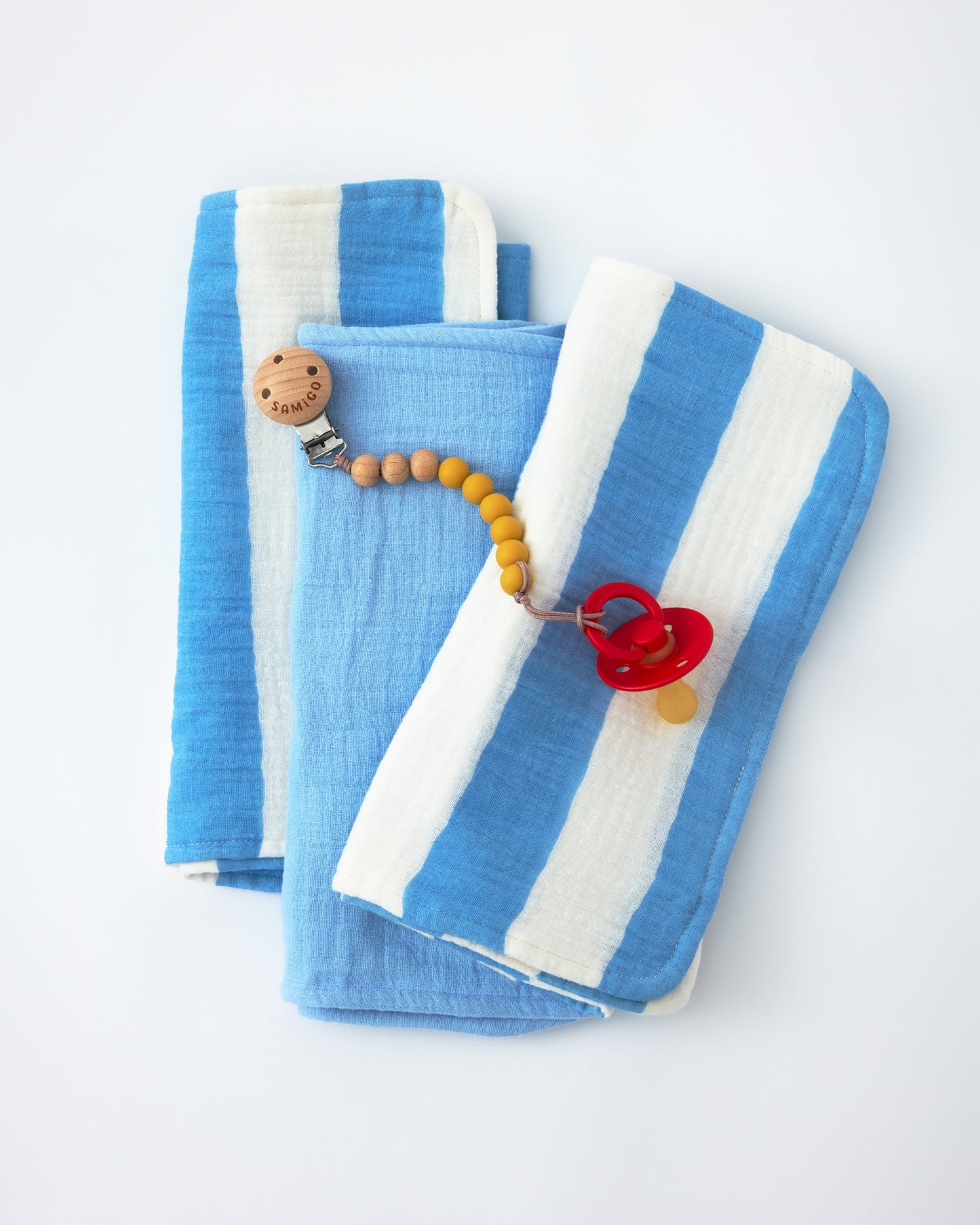 Organic Muslin Burp Cloth (Three-Pack) in Blue Wide Stripe and a pacifier clip on a beige background.