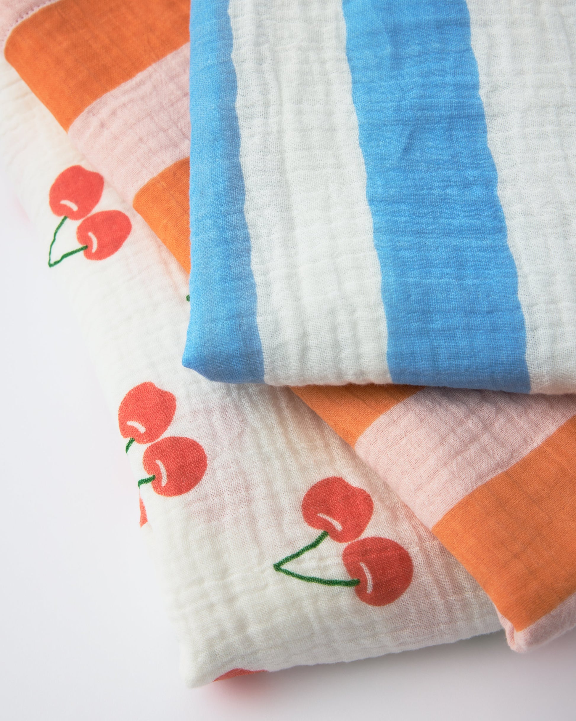 Three patterns of Organic Muslin Burp Cloth (Three-Pack) stacked, Blue Wide Stripe, Pink Wide Stripe, White Cherries, on a beige background.