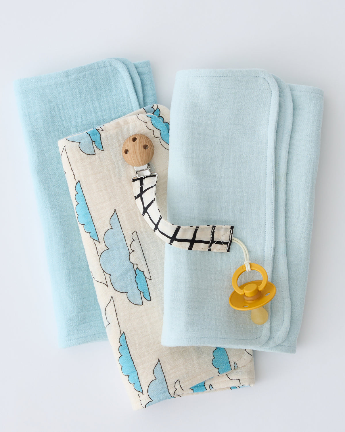 Organic Muslin Burp Cloth (Three-Pack) in Ivory Clouds, folded and stacked, with a Pacifier Clip in Black Grid, on a beige background.