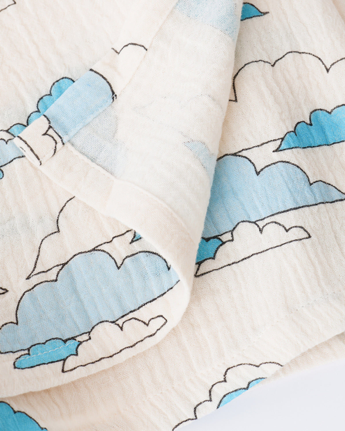Close up of the Organic Muslin Swaddle in Ivory Clouds, folded with corner flipped back to show reverse side, on a beige background.