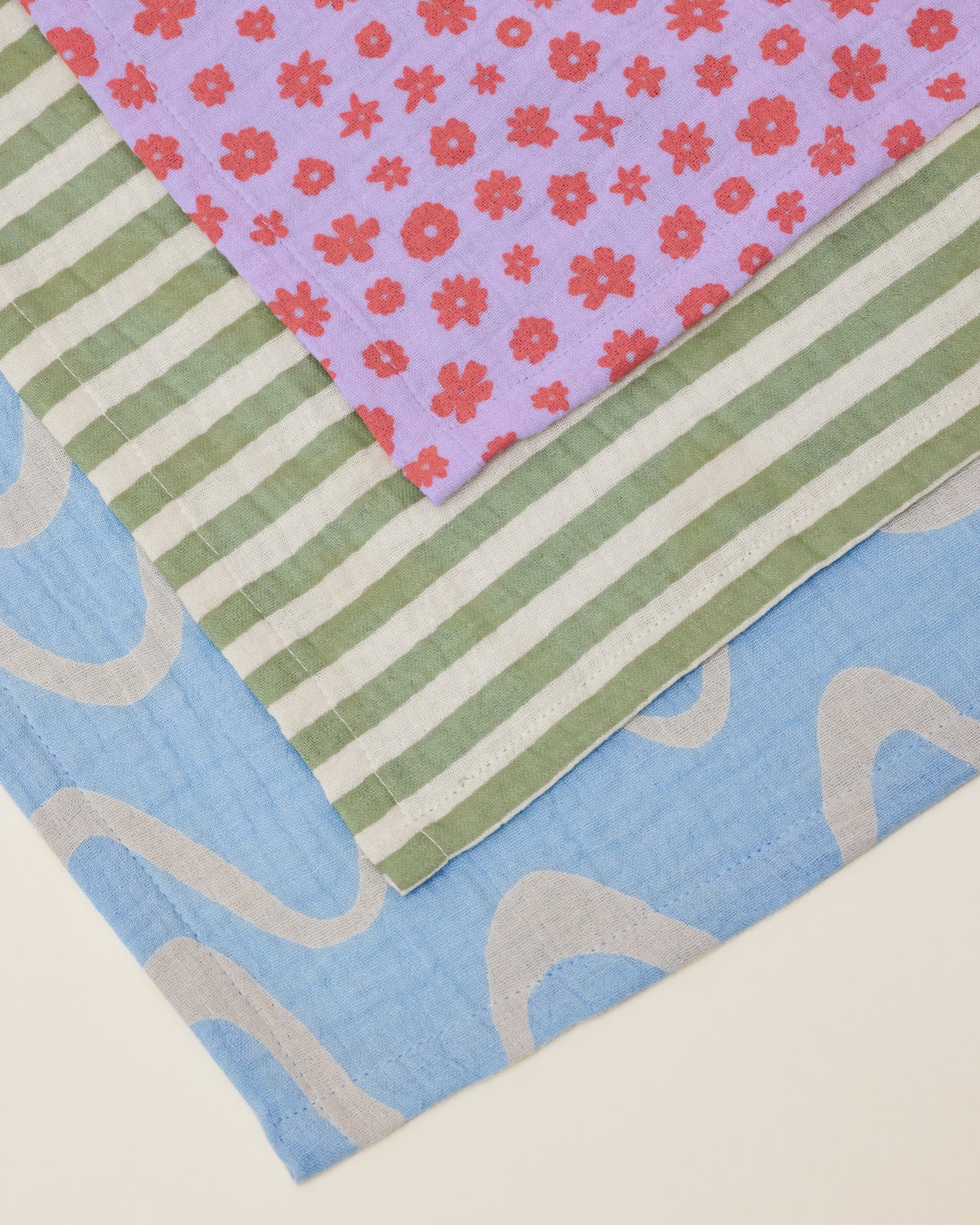 Close up of a stack of Organic Muslin Swaddle in the Lavender Cosmos, Sage Stripe and Blue Waves prints.