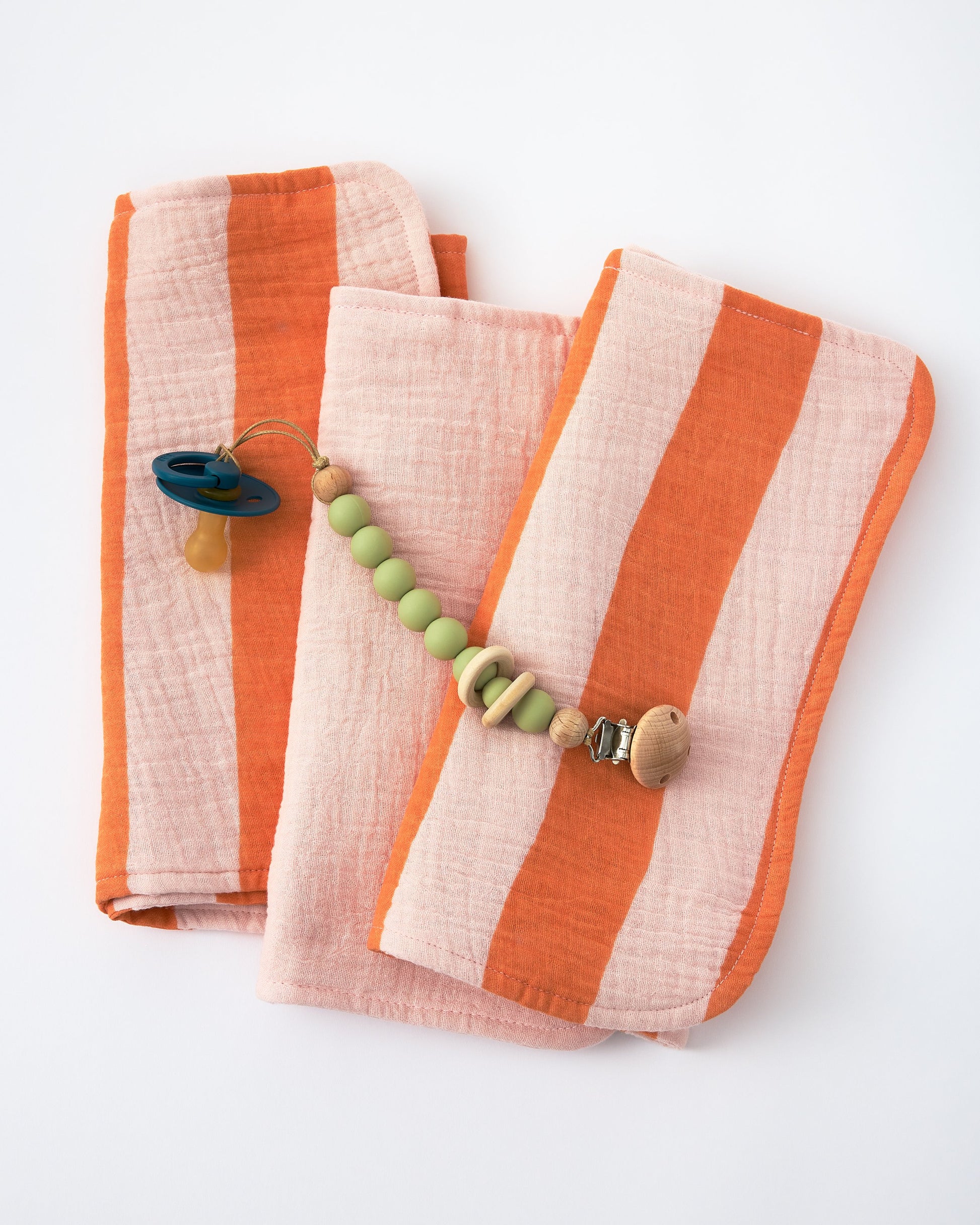 Organic Muslin Burp Cloth (Three-Pack) in Pink Wide Stripe with pacifier clip, on a beige background.