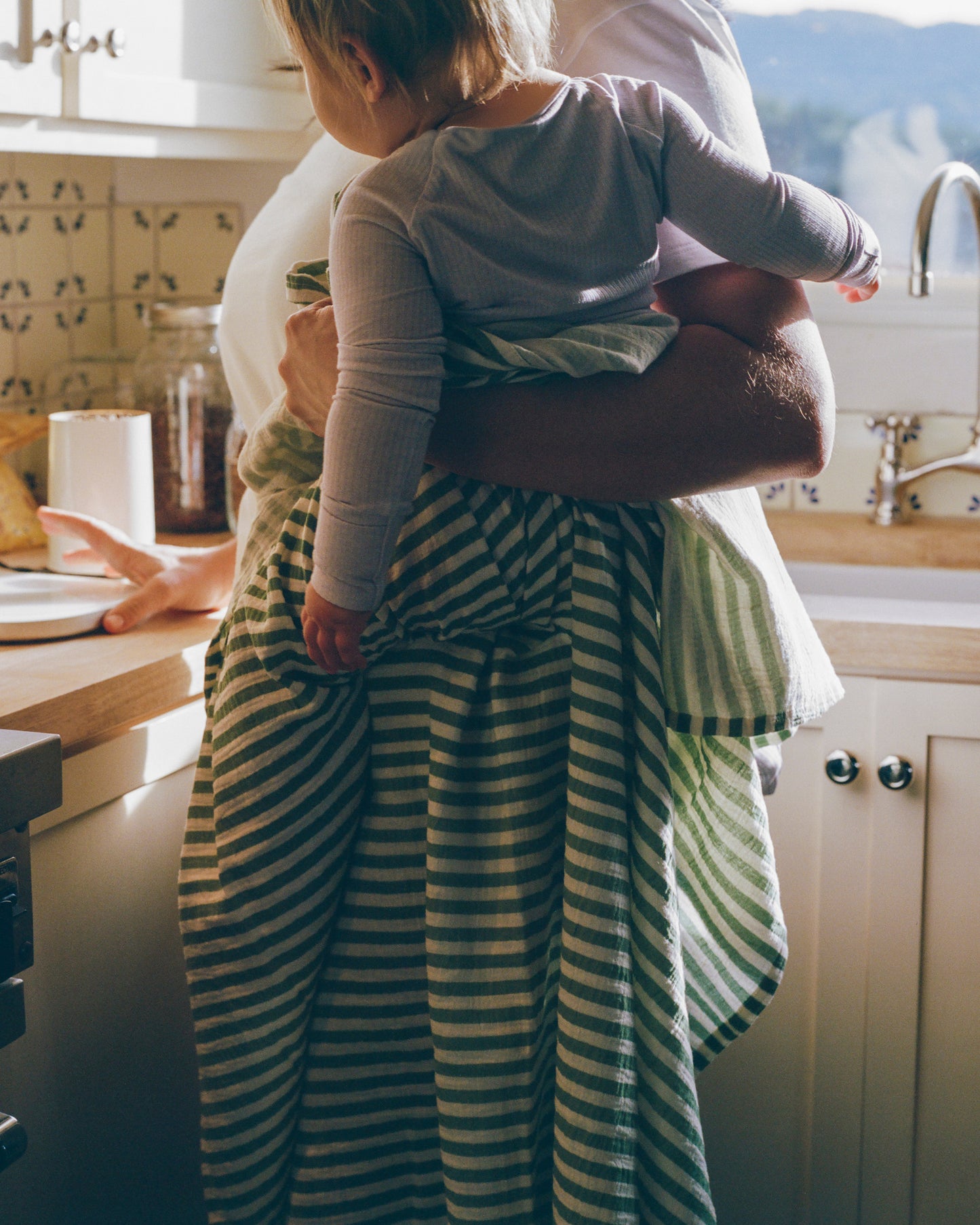 Person holding a child wearing the Ribbed Bamboo Baby Pajama and wrapped in Organic Muslin Swaddle, in a kitchen setting