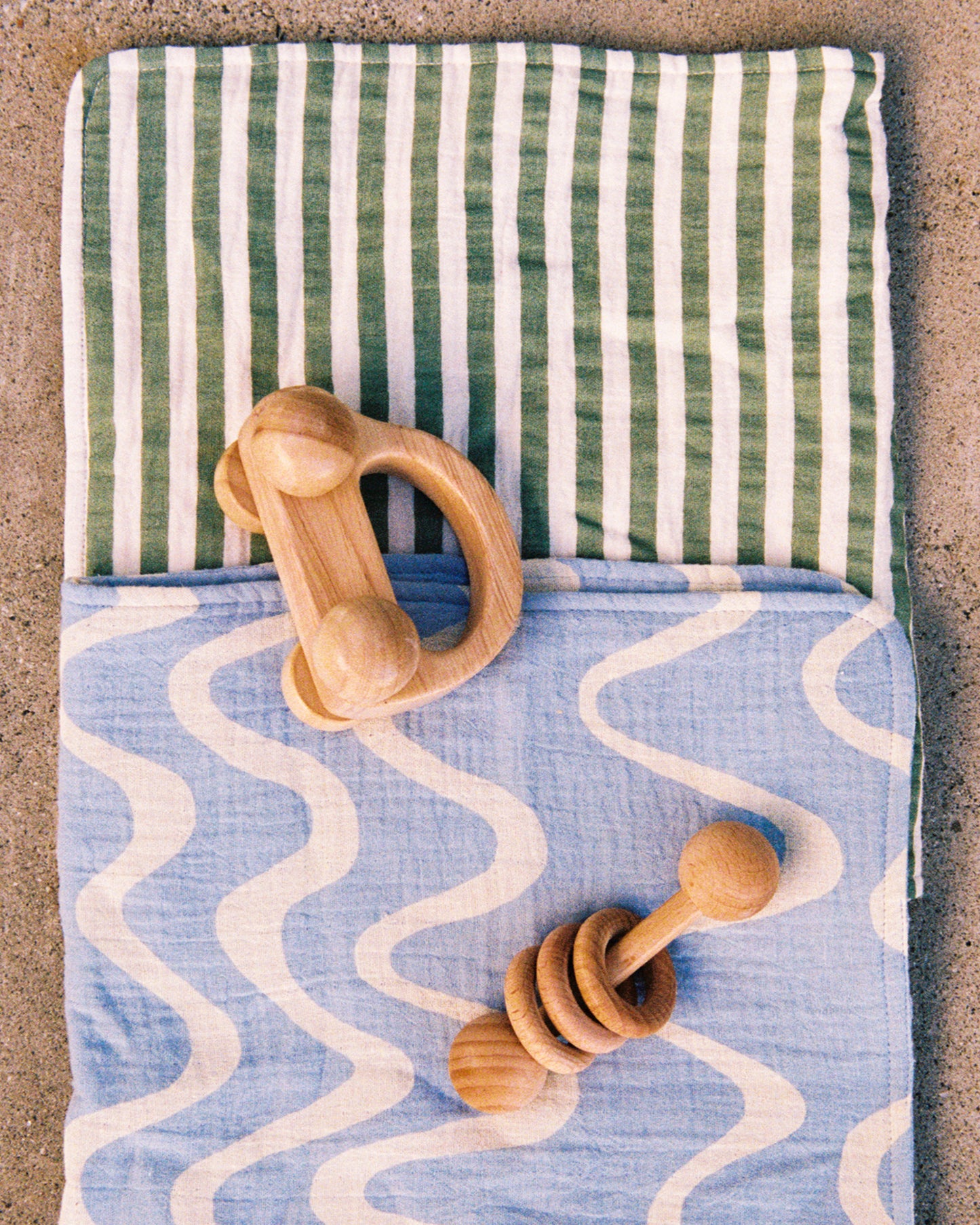 Two of the Organic Muslin Burp Cloth in the Sage Stripe and Blue Waves print, displayed with a wooden toy on a cement surface.