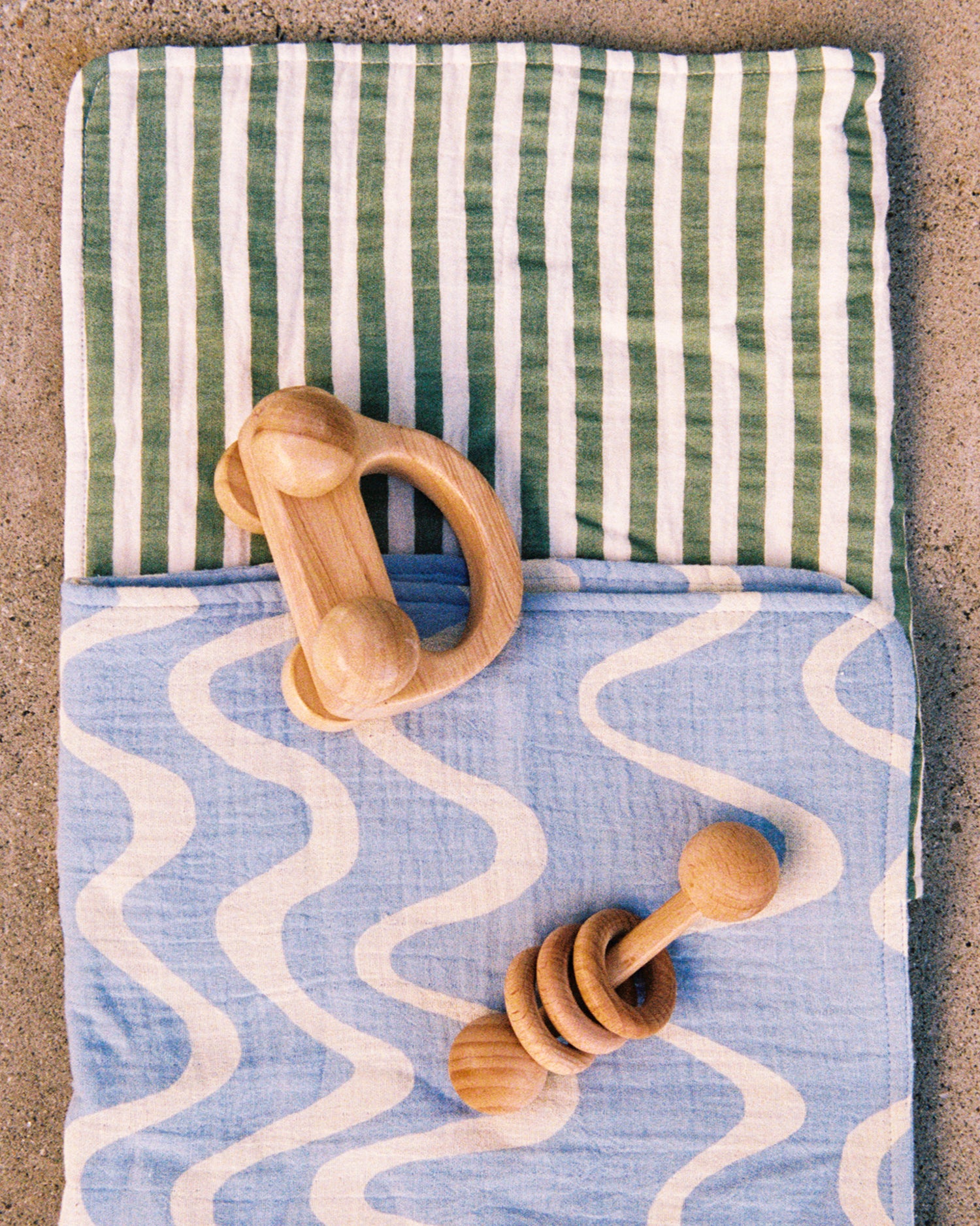 Two of the Organic Muslin Burp Cloth in the Sage Stripe and Blue Waves print, displayed with a wooden toy on a cement surface.