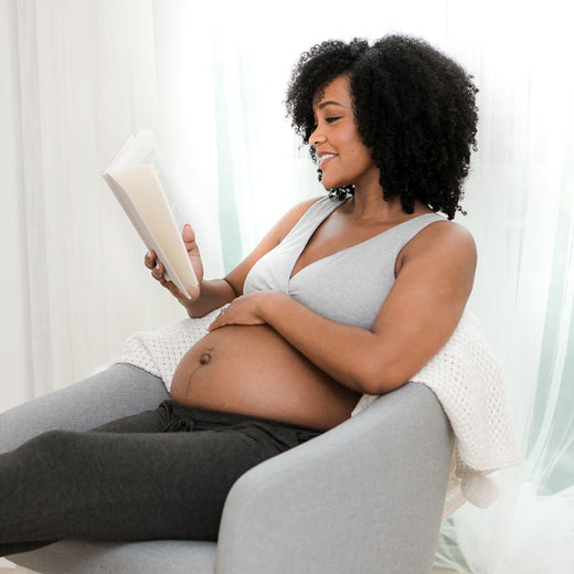 Pregnant woman sitting in a chair and reading a book, wearing a french terry racerback bra and lounge joggers.